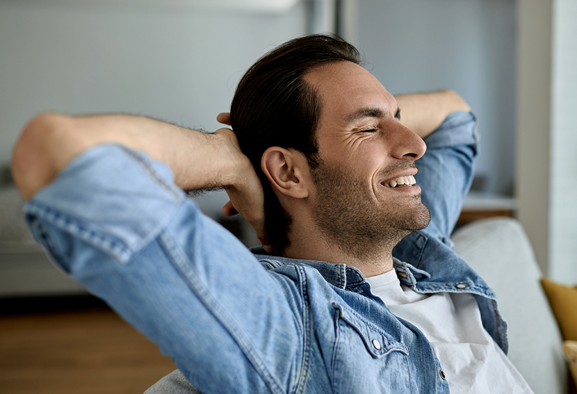 Man relaxing indoors with hands behind head, wearing blue denim shirt, smiling contentedly.