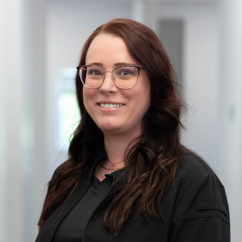 Woman with long brown hair and glasses smiling at camera in professional dental office setting.