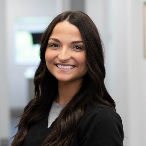 Young woman smiling at camera in professional dental office setting wearing black blazer.
