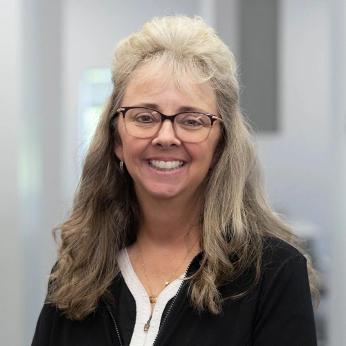 Professional woman with long hair and glasses smiling in dental office setting.