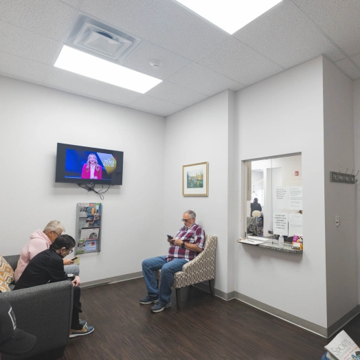 Two patients wait in modern dental office reception area with skylight and wall-mounted TV.