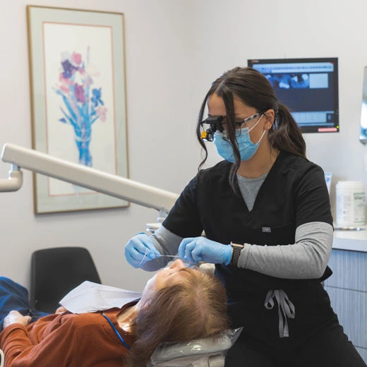 Dental hygienist wearing mask and gloves examines patient in modern dental office treatment room.
