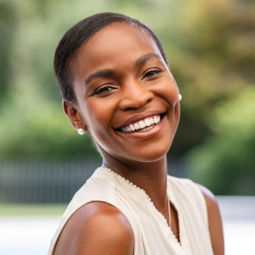 Woman smiling brightly outdoors, wearing white sleeveless top, dental care representation.
