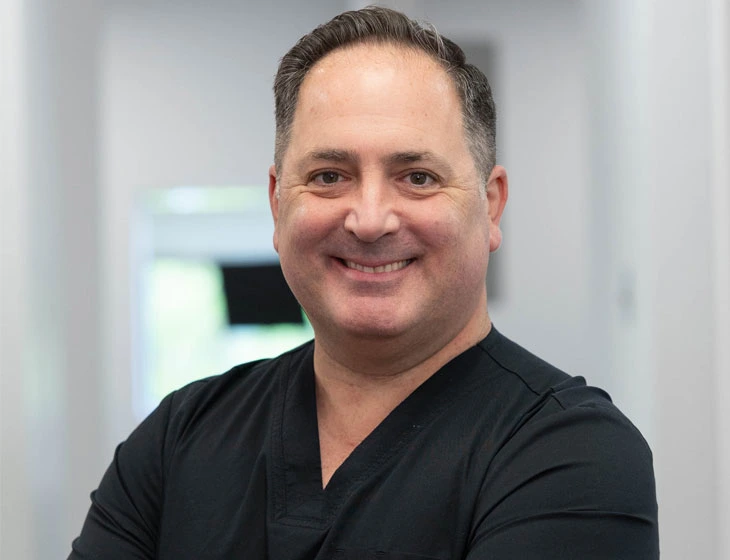 Male dentist in black scrubs smiling at camera in dental office setting.